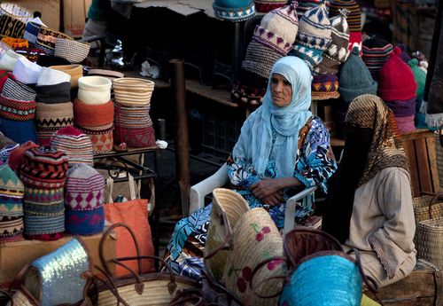 A vendor sitting among stacked knitwear and baskets at an indoor market stall.